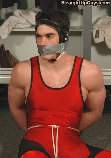 A muscular man wearing a red wrestling singlet and headgear sits bound and gagged with duct tape in a locker room setting.