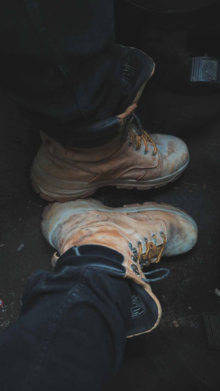 A pair of dirty tan work boots are worn with dark denim jeans in the footwell of a vehicle.