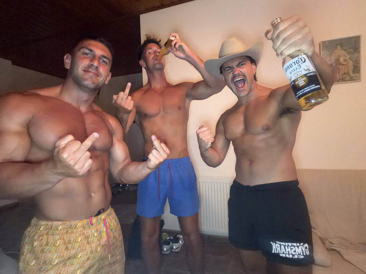 Three shirtless, muscular men in gym shorts and swim trunks drink beer and flex their physiques indoors.