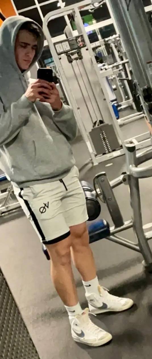 A young man in a grey hoodie and short gym shorts takes a mirror selfie inside a gym.