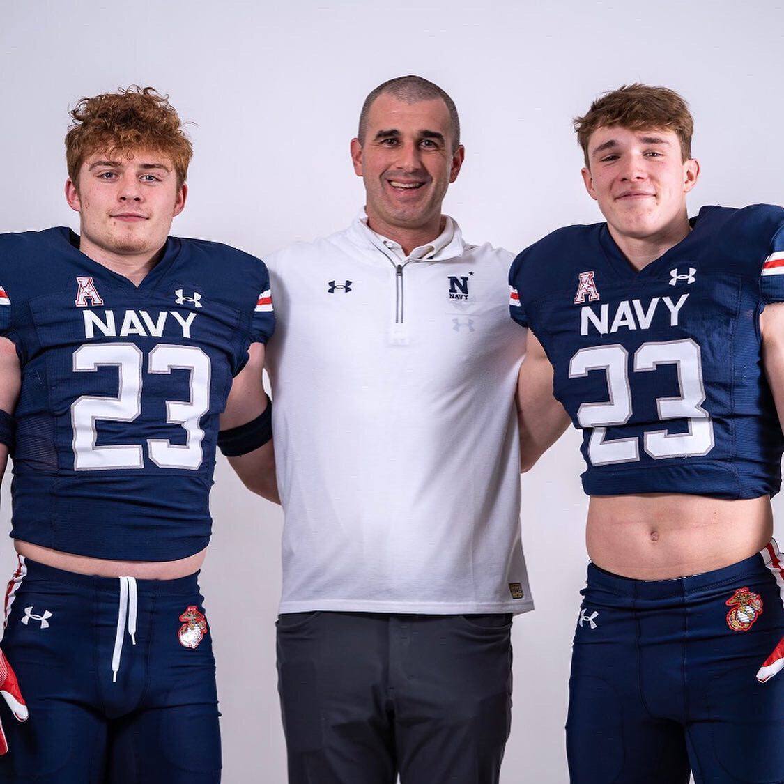 A mature coach stands with his arms around two young football players wearing padded Navy jerseys and tight athletic pants.
