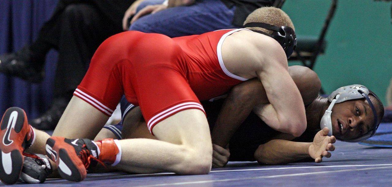 Two athletic wrestlers in singlets and headgear grapple on a blue mat, with one athlete in a red singlet pinning the other down.