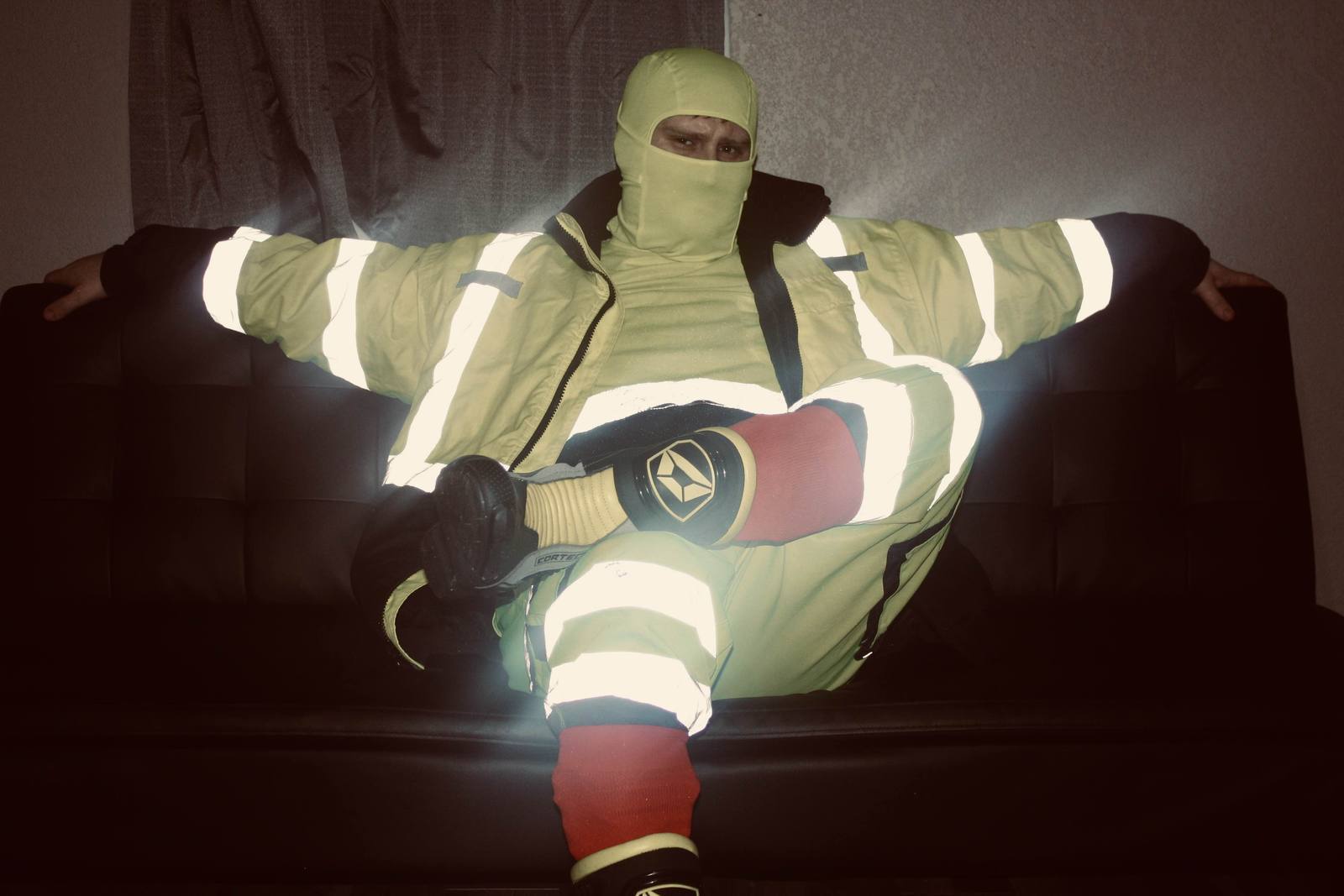 A man sits on a dark couch wearing a full high-visibility workwear uniform and a matching yellow balaclava.