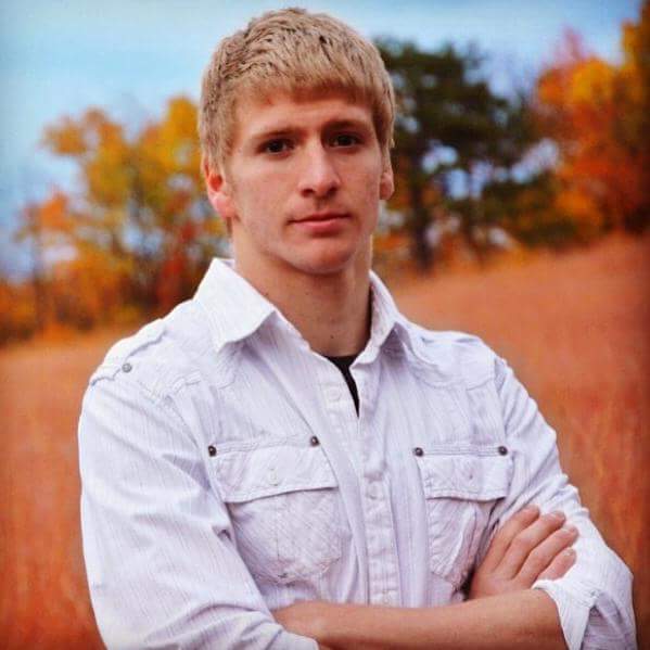 A young blonde man with his arms crossed poses outdoors in front of autumn foliage wearing a white button-down shirt.
