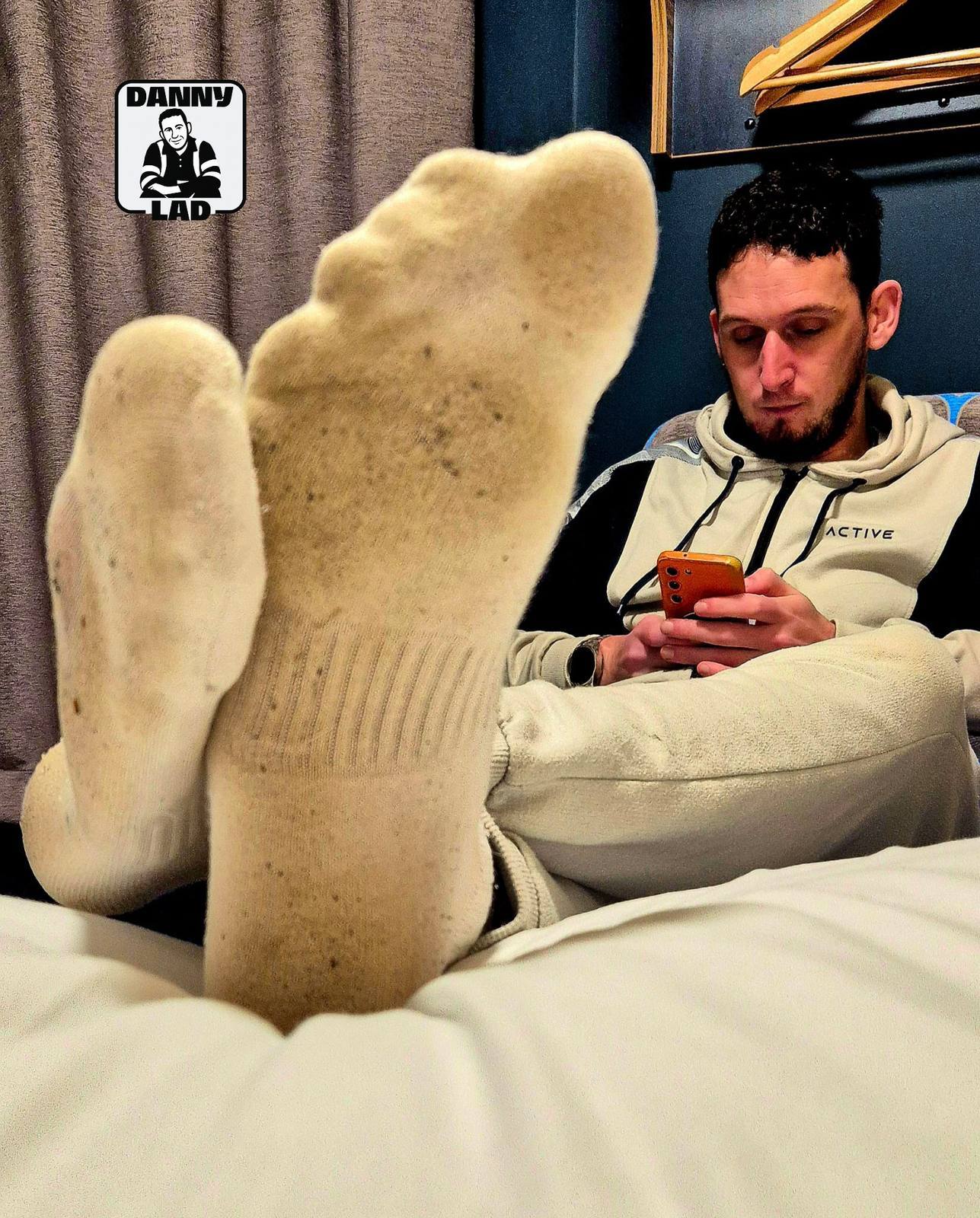A bearded man wearing a light-colored tracksuit sits on a bed with his dirty white socks prominently displayed in the foreground.