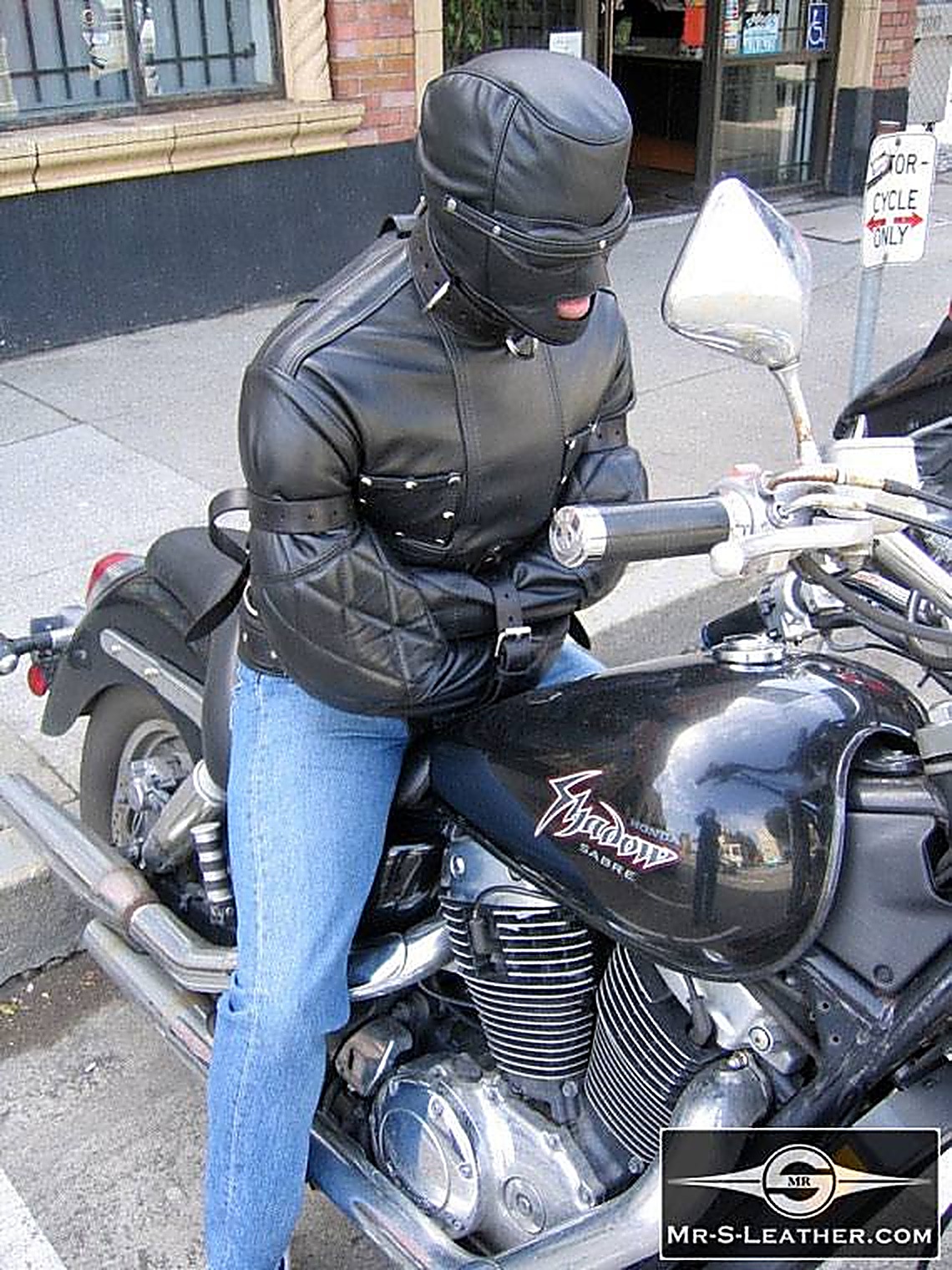 A person clad in a black leather straitjacket and matching leather hood sits restricted atop a parked motorcycle outdoors.