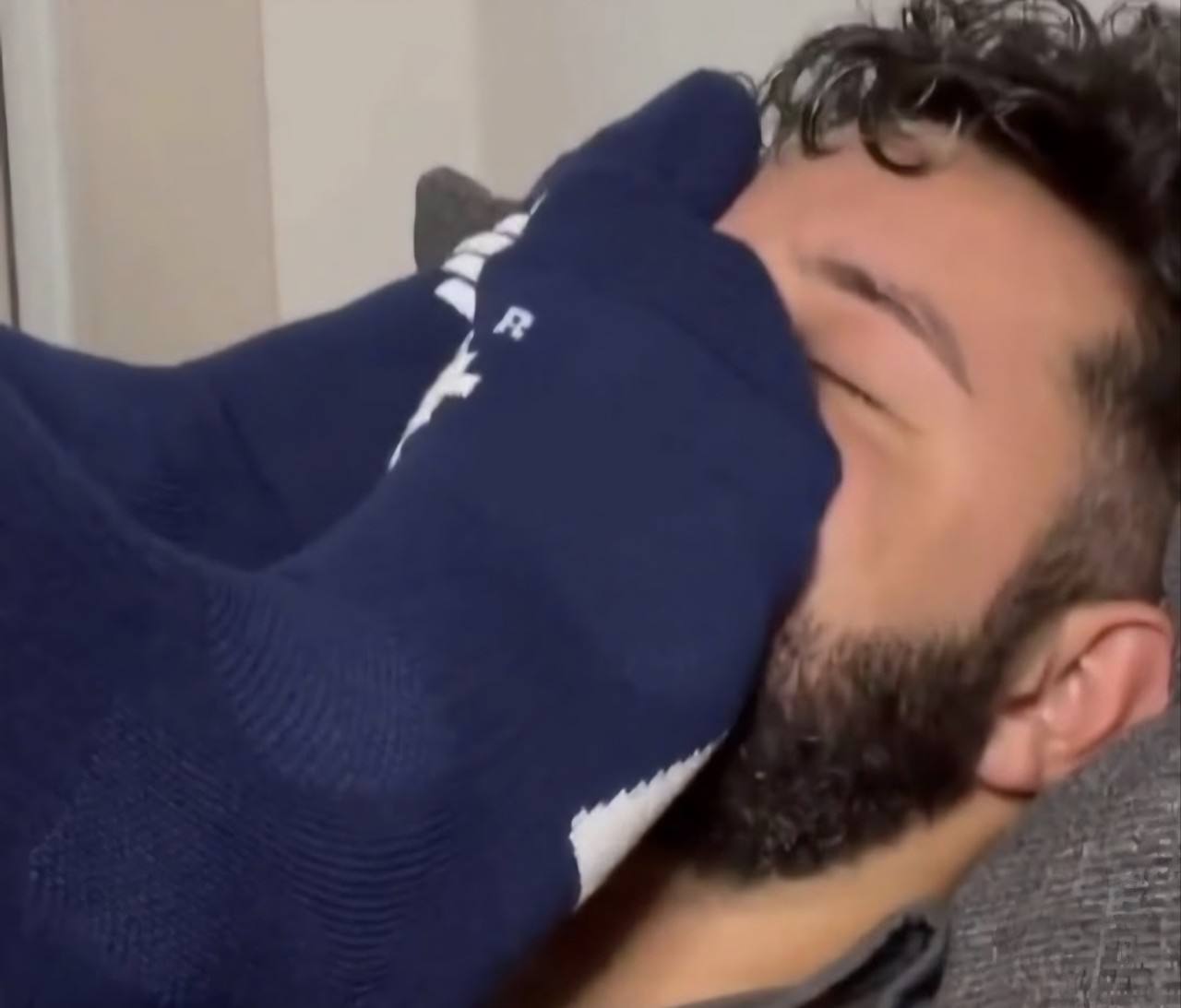 A bearded man lies back while a foot wearing a blue athletic sock is pressed firmly against his face.