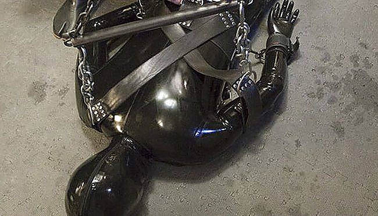 A person clad in a full black latex gimp suit and hood is bound on the floor with heavy straps, chains, cuffs, and a metal spreader bar.
