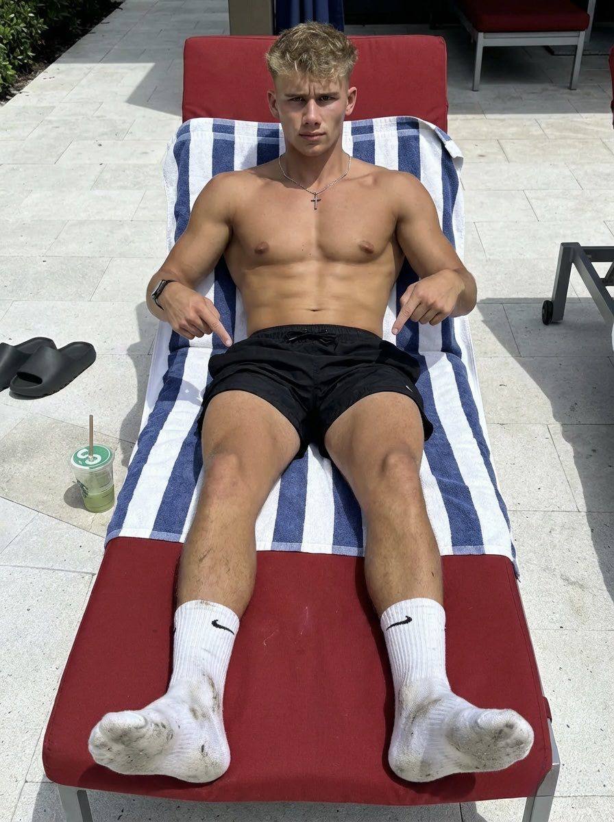 A shirtless, muscular young man in black gym shorts and soiled white crew socks reclines on a poolside lounge chair.