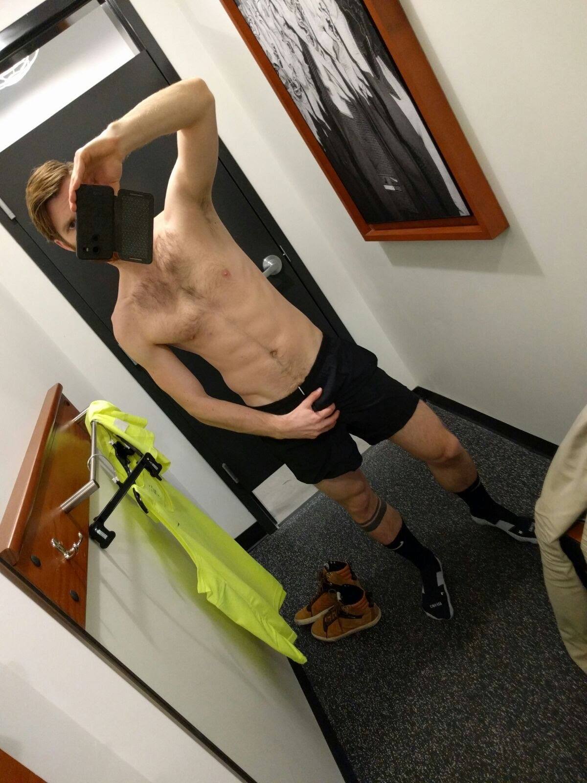 A shirtless, hairy-chested man with visible abs poses for a mirror selfie in black gym shorts and socks inside a changing room.