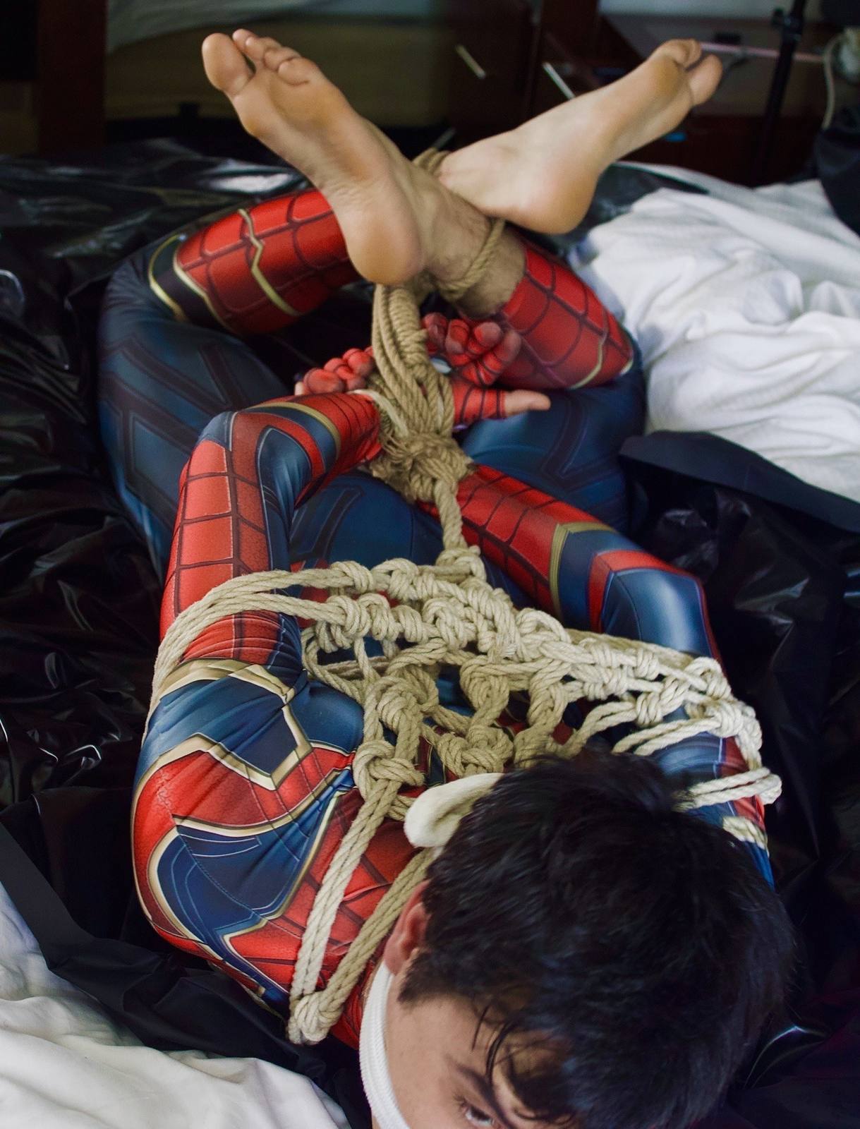 A man in a Spidey-themed spandex suit is bound in complex rope bondage and gagged while lying on a bed.