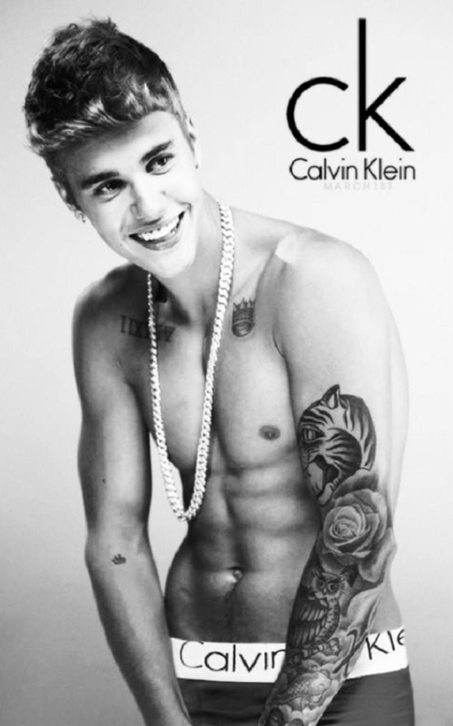A shirtless tattooed young man poses in black and white wearing Calvin Klein underwear and a silver chain necklace.