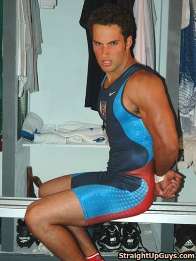 A muscular man in a tight red, blue, and white Nike wrestling singlet sits on a locker room bench with his hands behind his back.