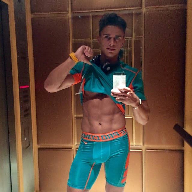 A fit young man in teal and orange compression gear pulls up his shirt to reveal defined abs while taking a mirror selfie in an elevator.