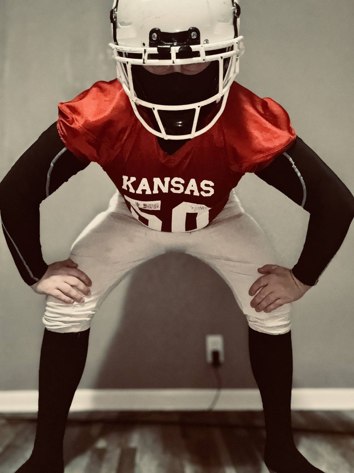 A man in a full American football uniform, including a white helmet and red Kansas jersey, poses in a set stance indoors.