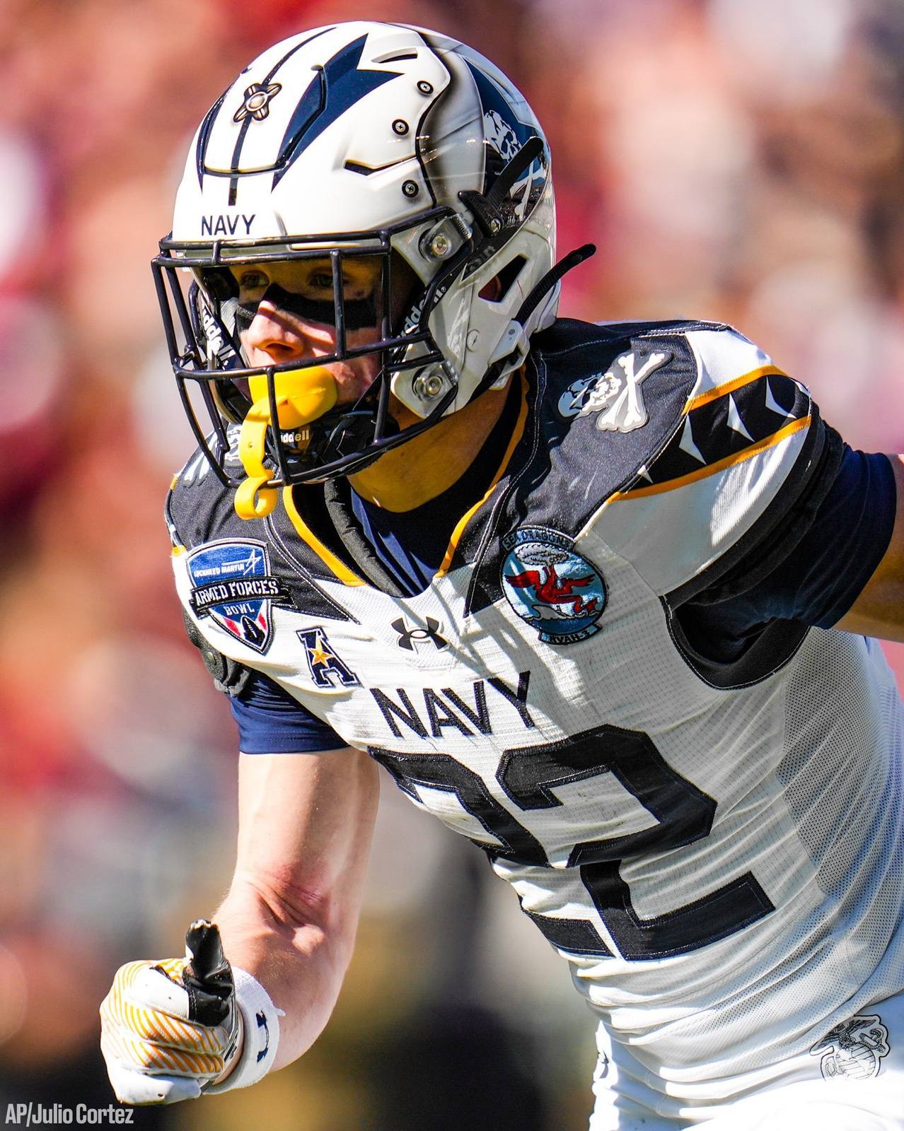 A young football player in a full Navy uniform, including a helmet with a yellow mouthguard and face mask, is captured in action during a game.