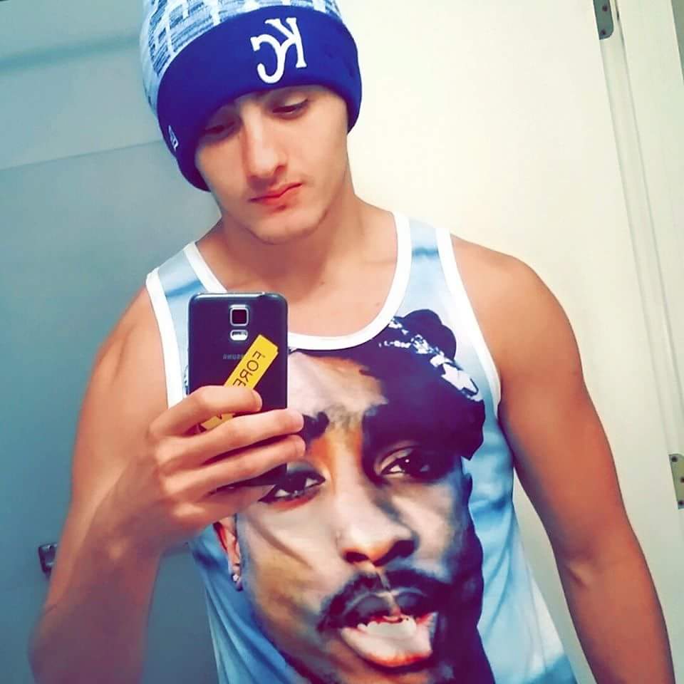 A young man wearing a beanie and a graphic tank top takes a mirror selfie indoors.