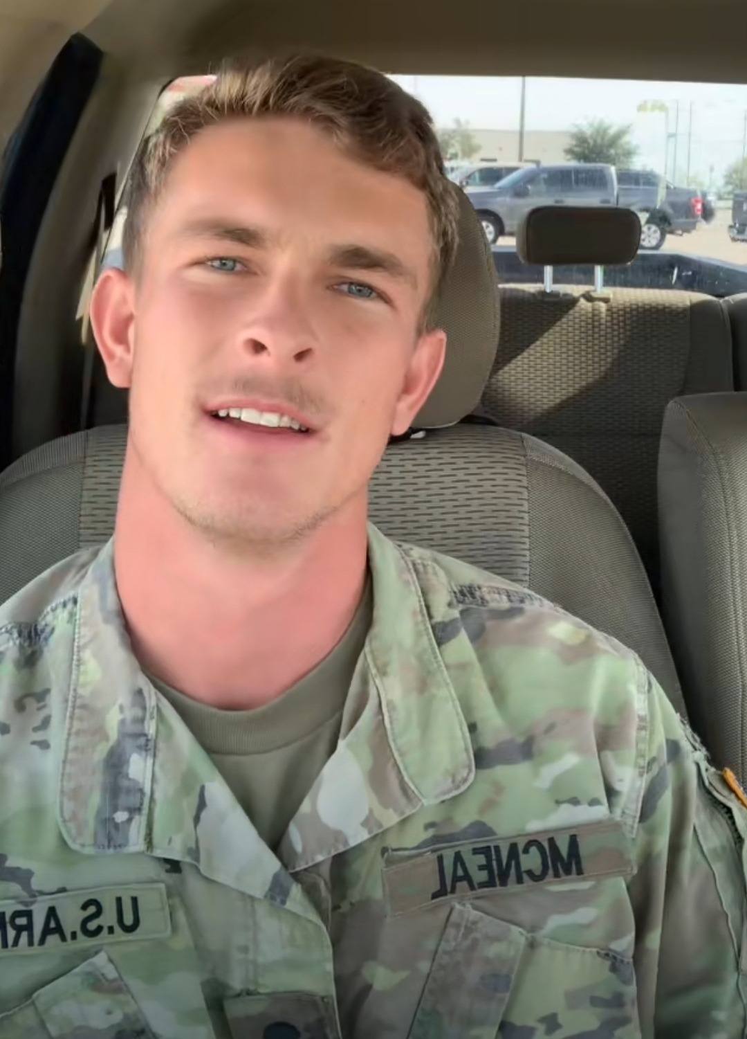 A young man wearing a US Army camouflage uniform sits in the driver's seat of a vehicle.