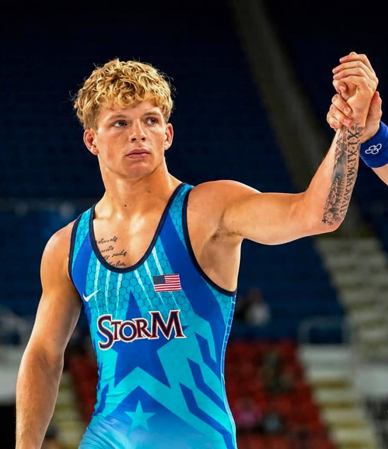A young athletic man with blond curly hair and tattoos wears a blue patterned wrestling singlet as his arm is raised in a wrestling arena.