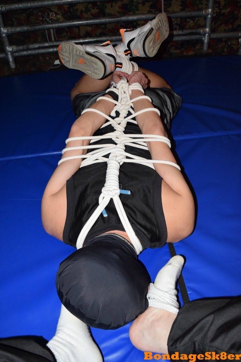A submissive male in a spandex hood and gym gear is hogtied with white rope on a blue mat.