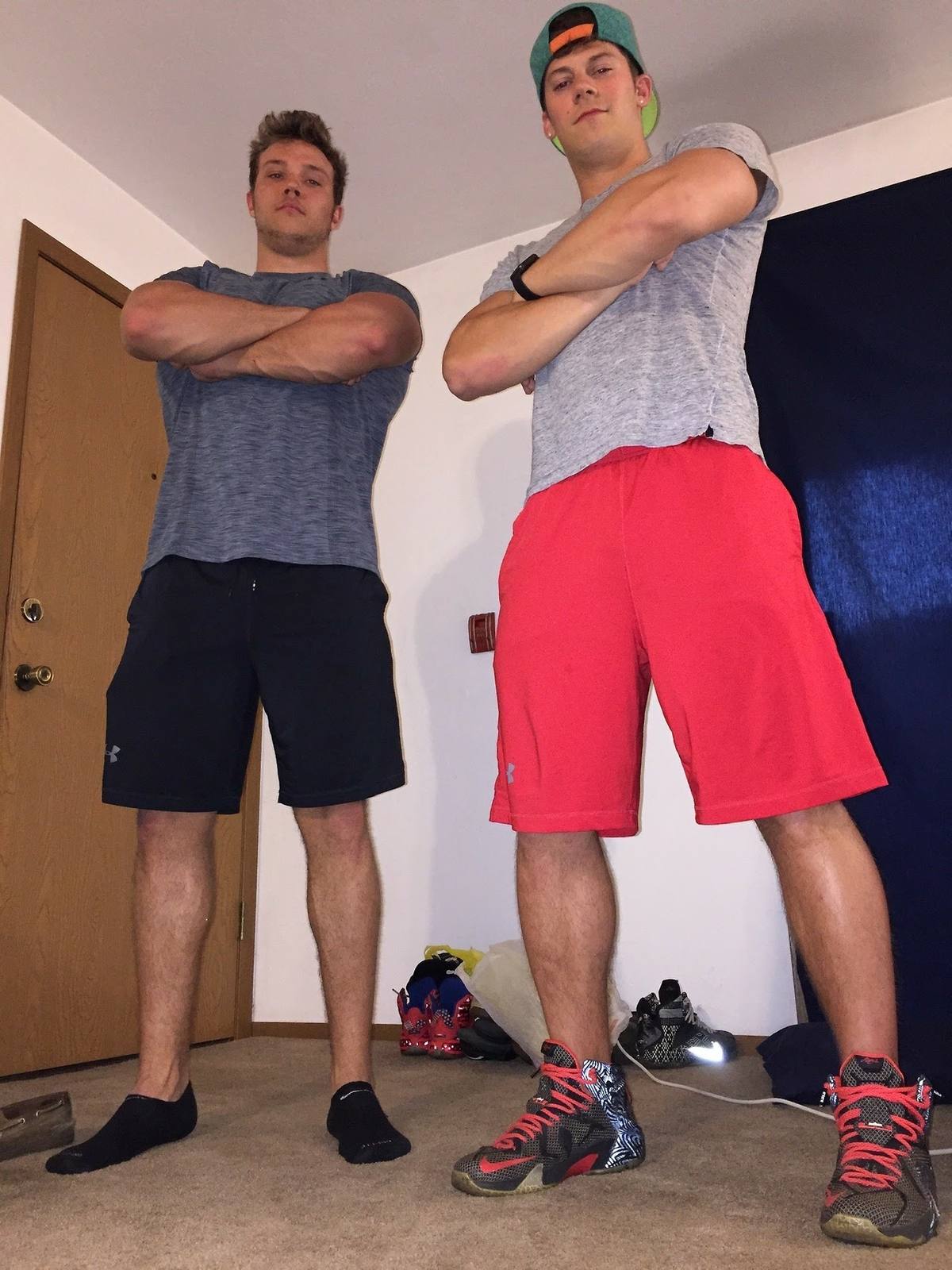 Two athletic young men in gym shorts and t-shirts stand side-by-side with arms crossed.