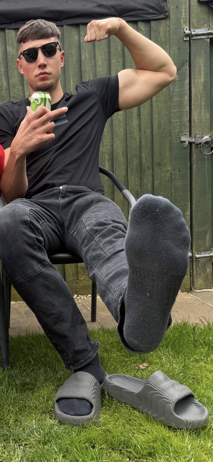 A young man sits outdoors, flexing his bicep and prominently displaying his feet in socks.
