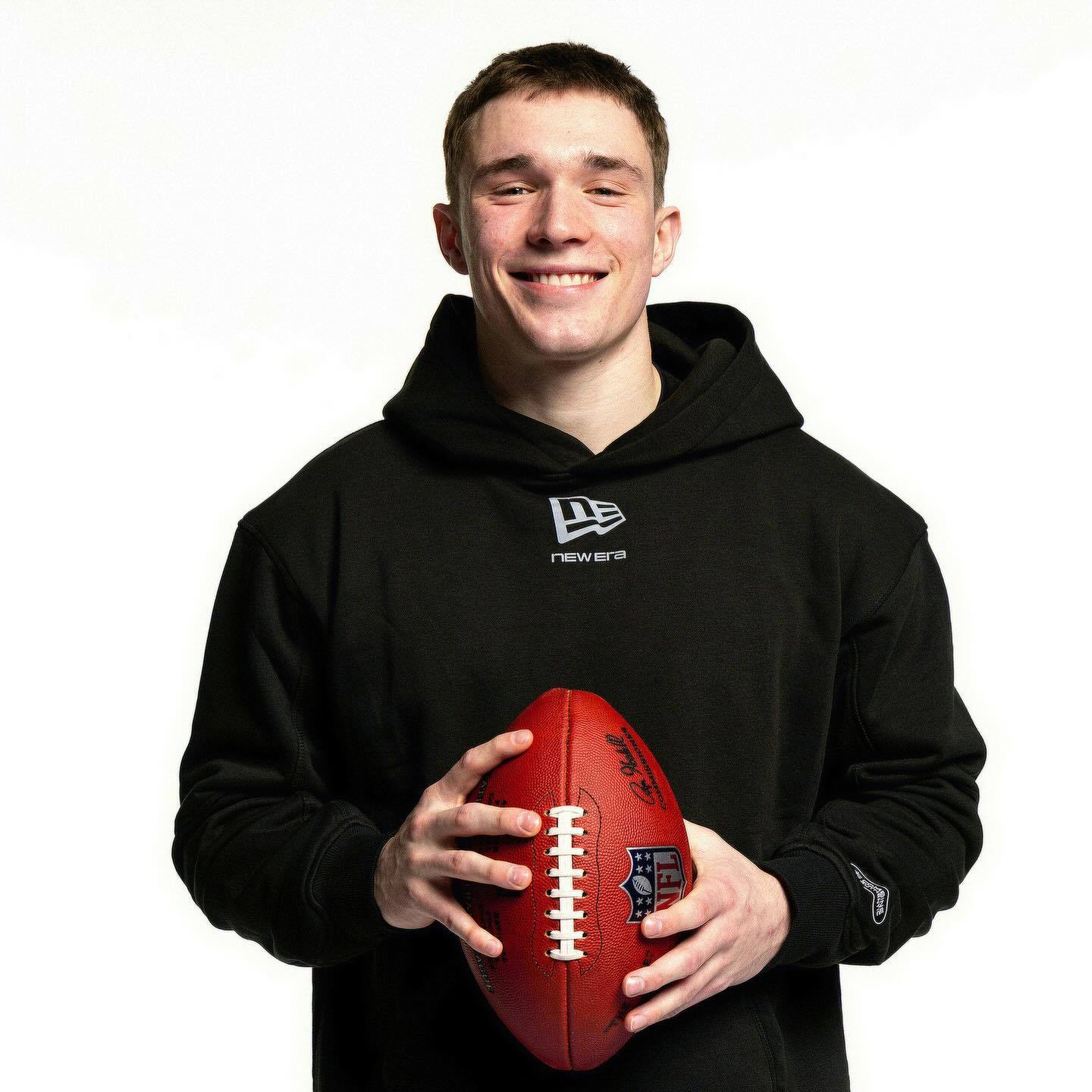 A smiling young man with a short haircut wears a black New Era hoodie while holding an NFL football against a white studio background.