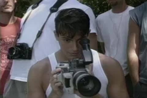 A young man wearing a white tank top operates a video camera while surrounded by others outdoors.