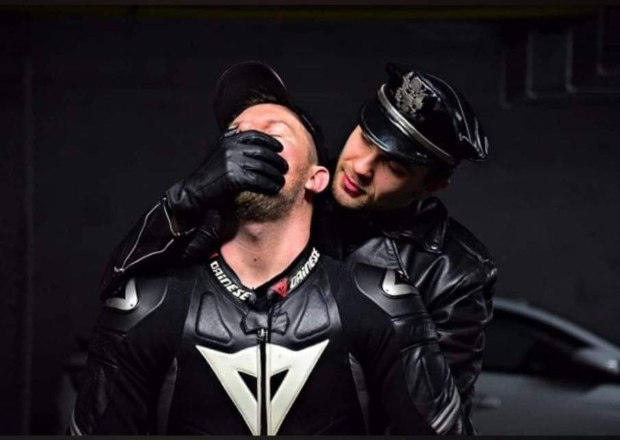 A man in leather gear and a muir cap holds a gloved hand over the mouth of another man wearing a Dainese motorcycle suit.