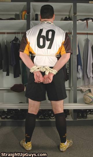 A man in a rugby uniform stands bound with rope behind his back in a locker room.