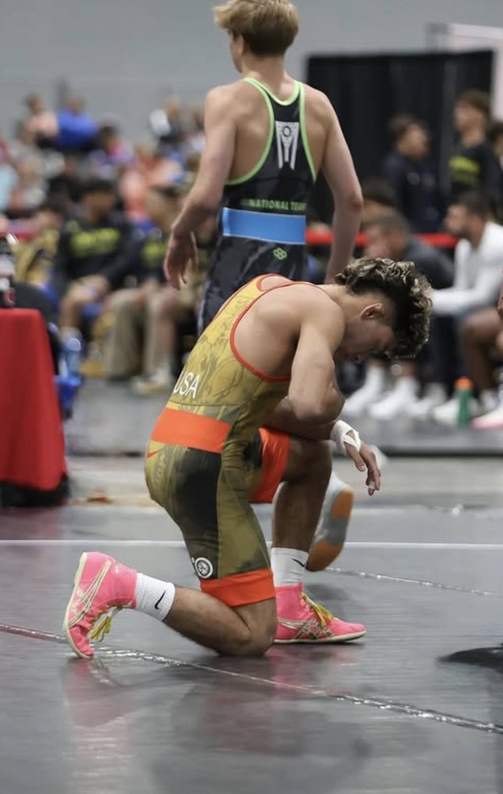 A young man wearing a patterned wrestling singlet and pink shoes kneels on a competition mat.