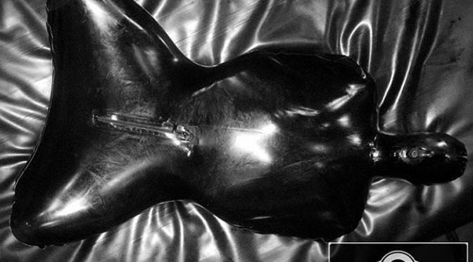 A figure is fully enclosed inside a shiny black inflatable latex suit lying on a glossy surface.