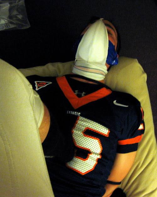 A man in a football jersey lies on his back with his hands bound behind him and an athletic sock pulled over his face as a makeshift hood.