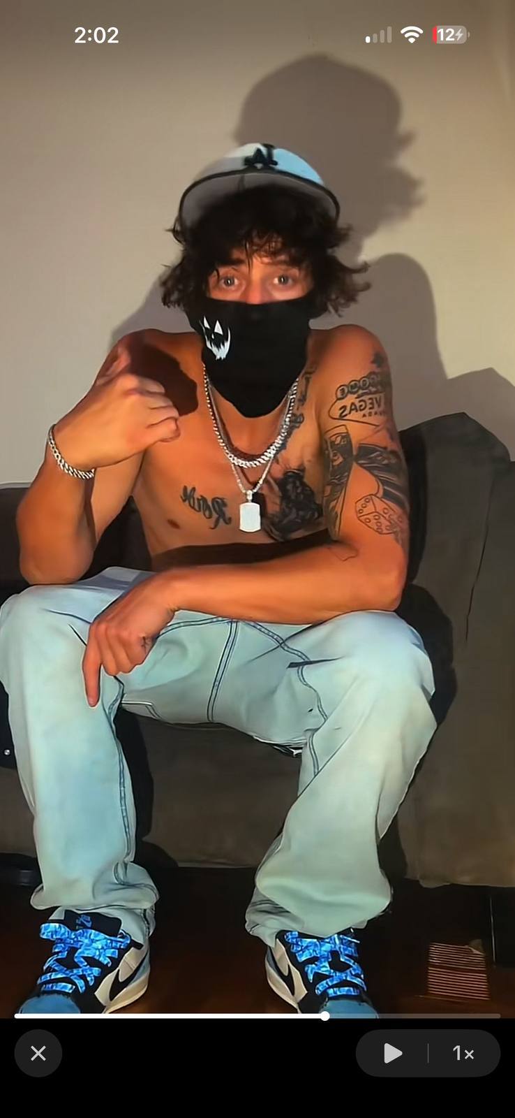 A shirtless, tattooed young man wearing a face mask and light blue jeans sits on a couch.