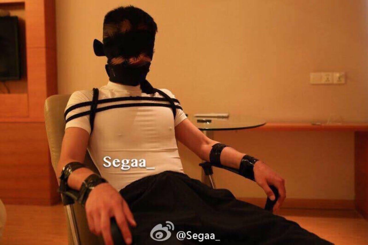 A young man is blindfolded, gagged, and restrained to an armchair in a hotel room with chest straps and tape.