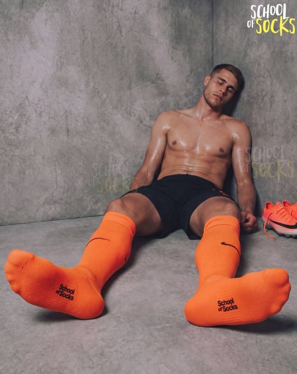 A sweaty, muscular young man in black gym shorts and bright orange football socks rests against a concrete wall.