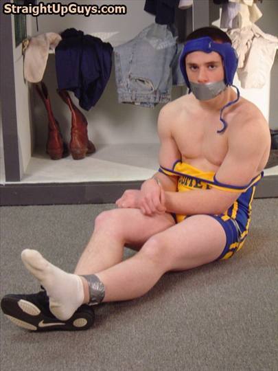 An athletic man in a wrestling singlet and headgear sits bound and gagged with duct tape on a locker room floor.