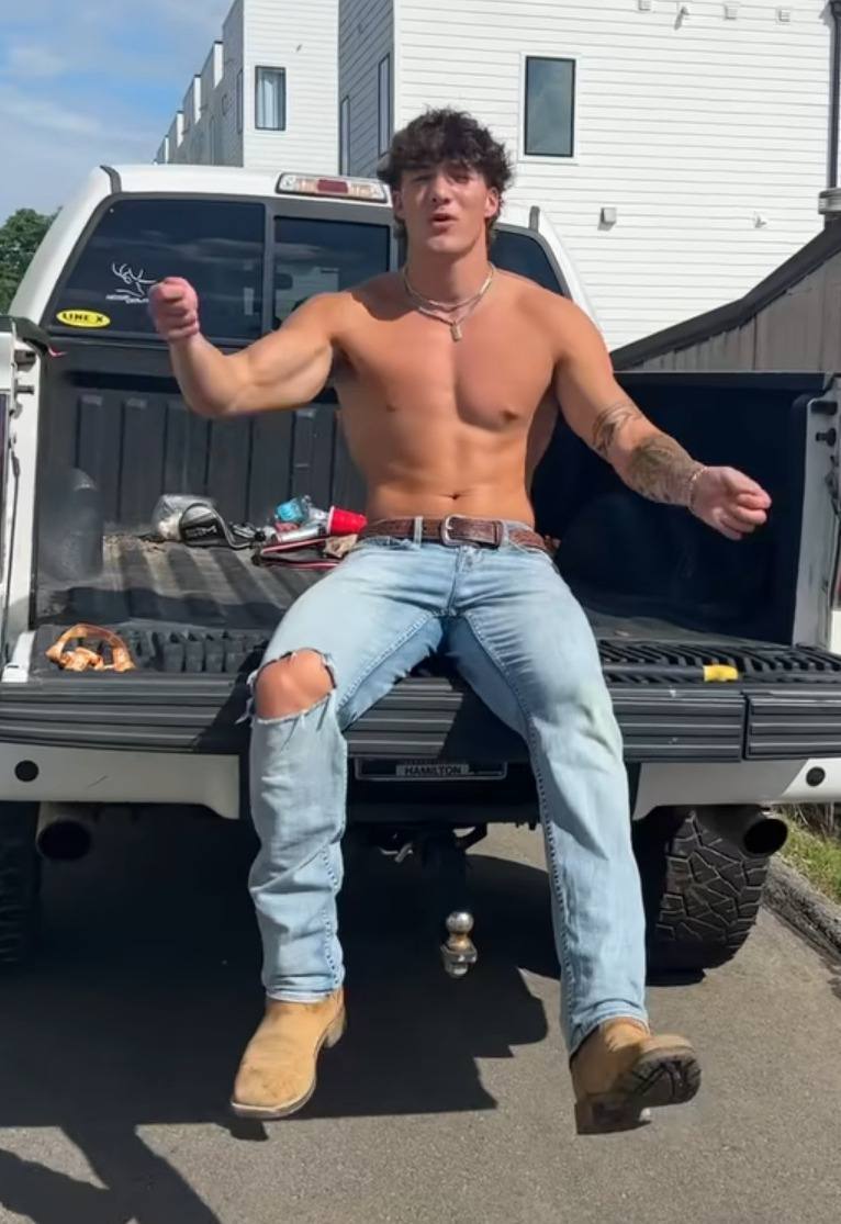 A shirtless muscular man in distressed jeans and boots sits on the tailgate of a pickup truck.
