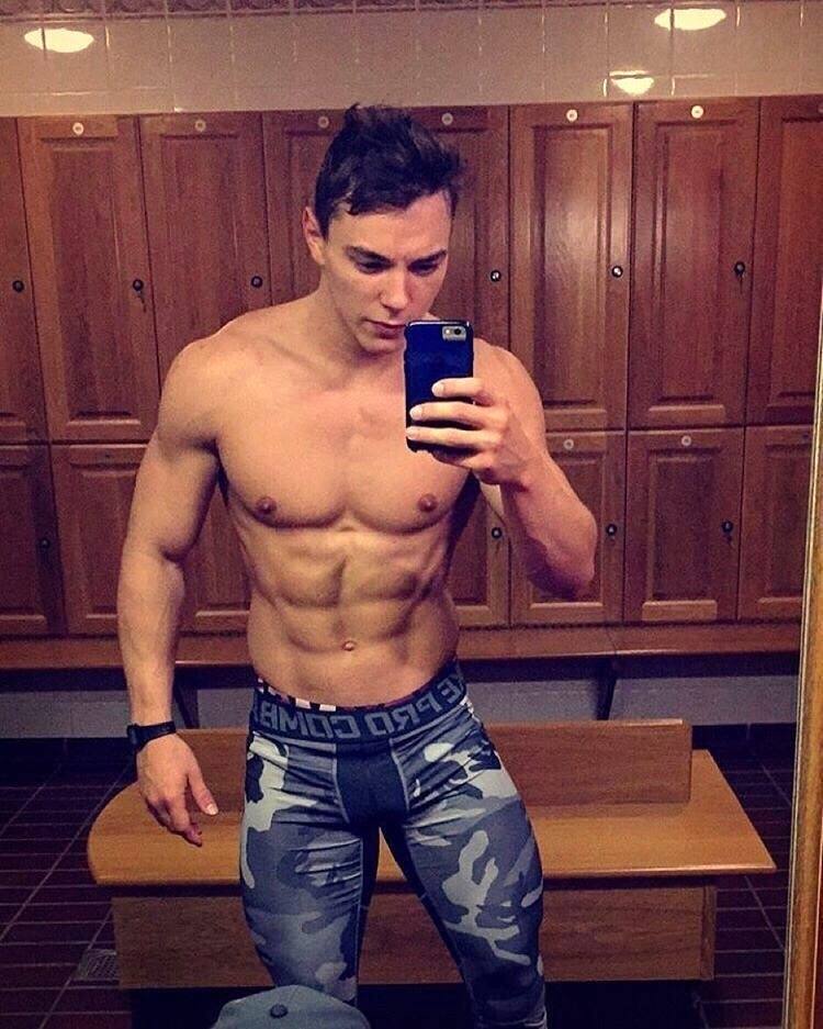 A muscular, shirtless man in camo-patterned compression leggings takes a mirror selfie in a gym locker room.