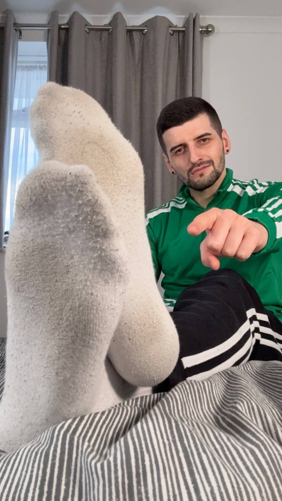 A bearded man in a green track jacket presents his white-socked feet to the camera while pointing at the viewer from a bed.
