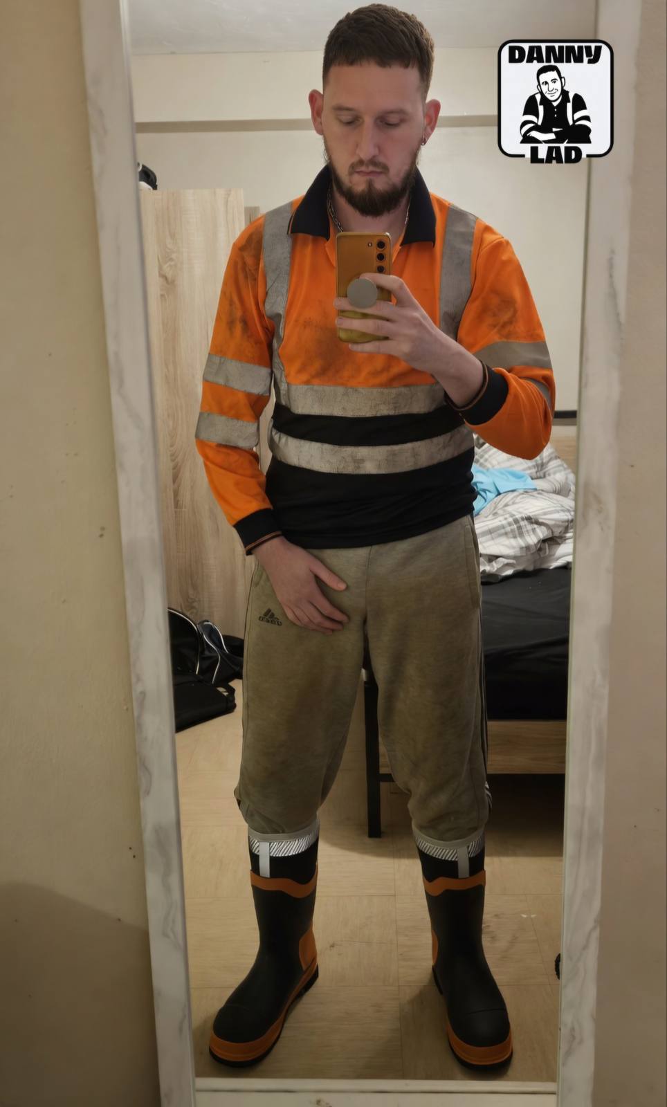 A bearded man takes a mirror selfie while wearing a dirty high-visibility work shirt, grey sweatpants, and rubber boots.