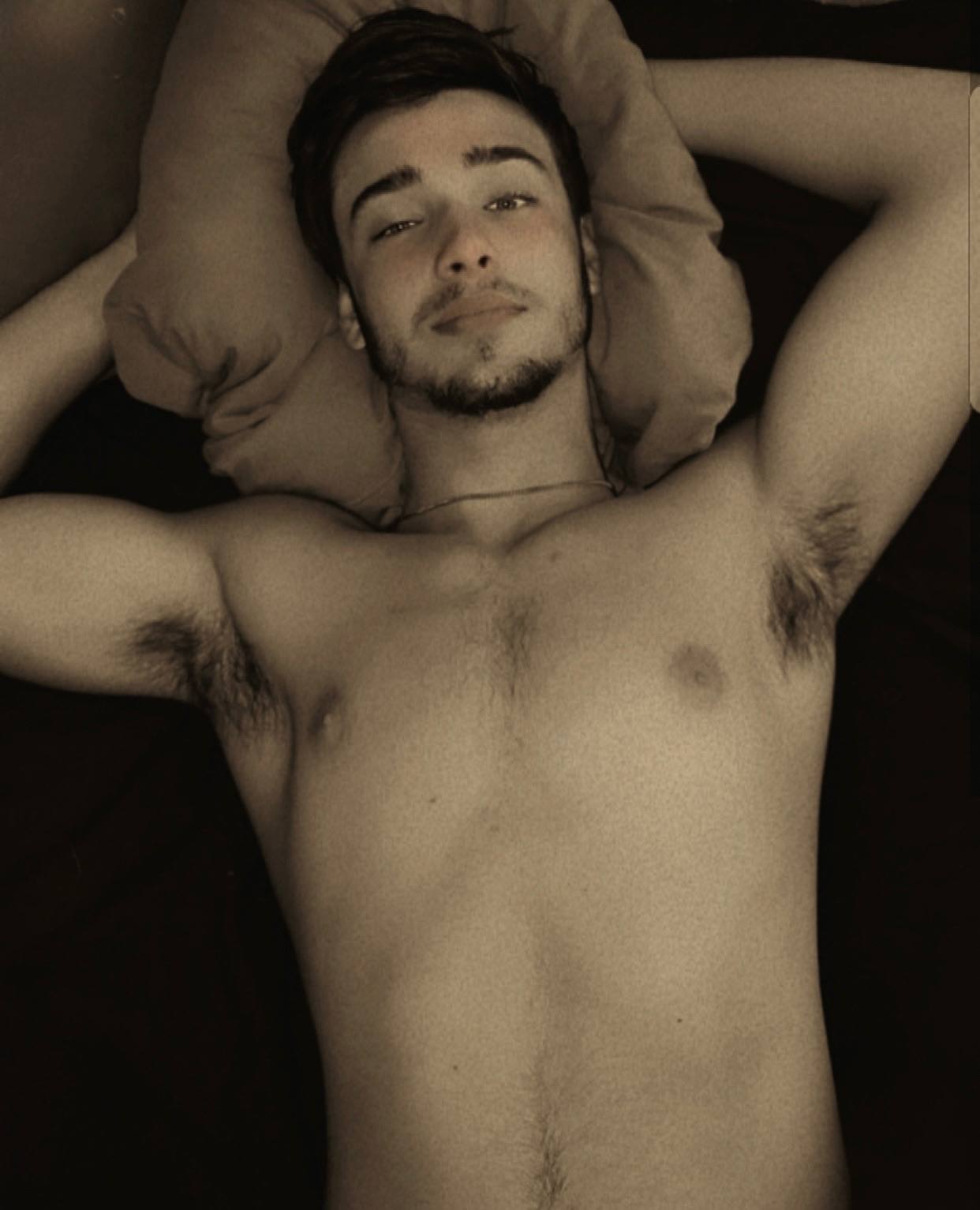 A shirtless twunk with a beard lies in bed with his hands behind his head, revealing hairy armpits.