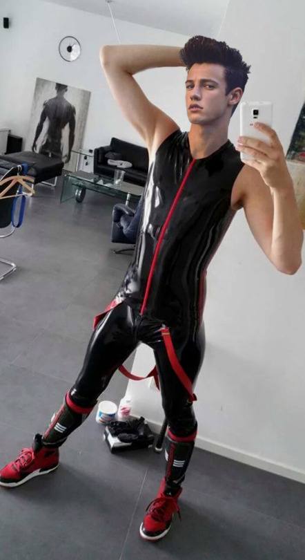 A young man in a black latex catsuit with red accents poses for a selfie indoors.