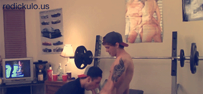 A shirtless twunk with a shoulder tattoo receives oral sex from another man in a room with a weight bench.
