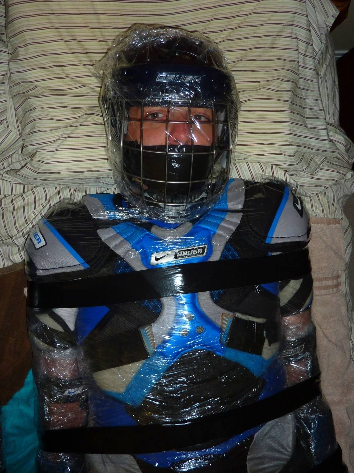 A man in full hockey gear is heavily bound with clear plastic wrap and black duct tape while his mouth is taped shut under a caged helmet.