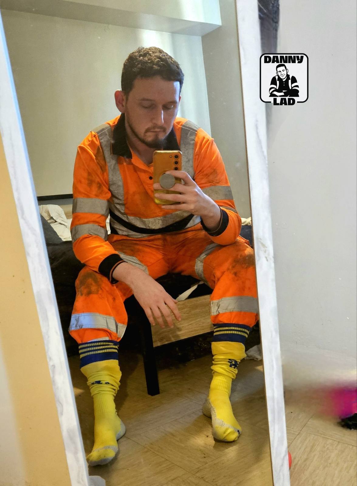 A bearded man wearing a dirty orange hi-vis boiler suit and long yellow football socks takes a mirror selfie in a bedroom.