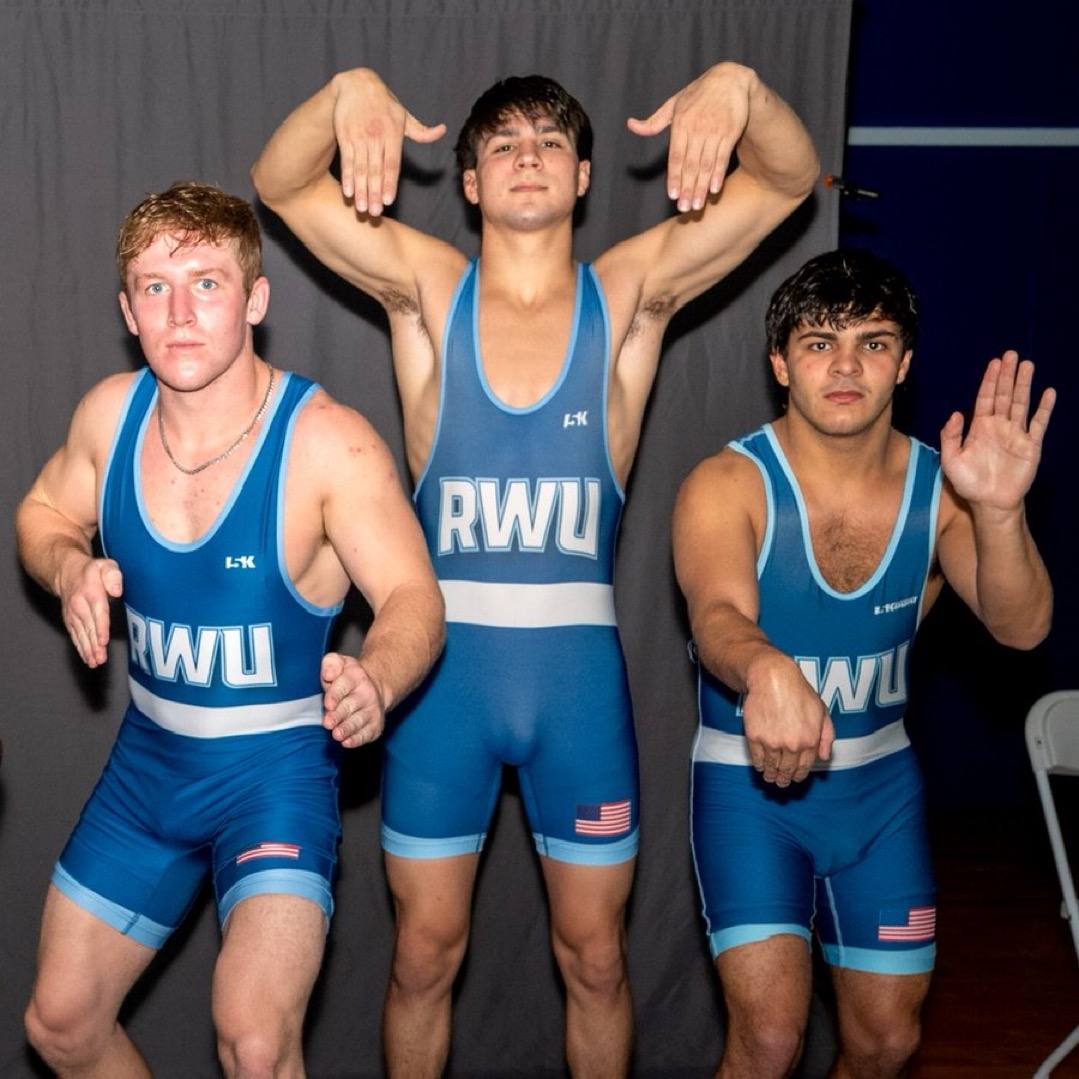 Three young men in blue wrestling singlets pose for a group photo indoors.