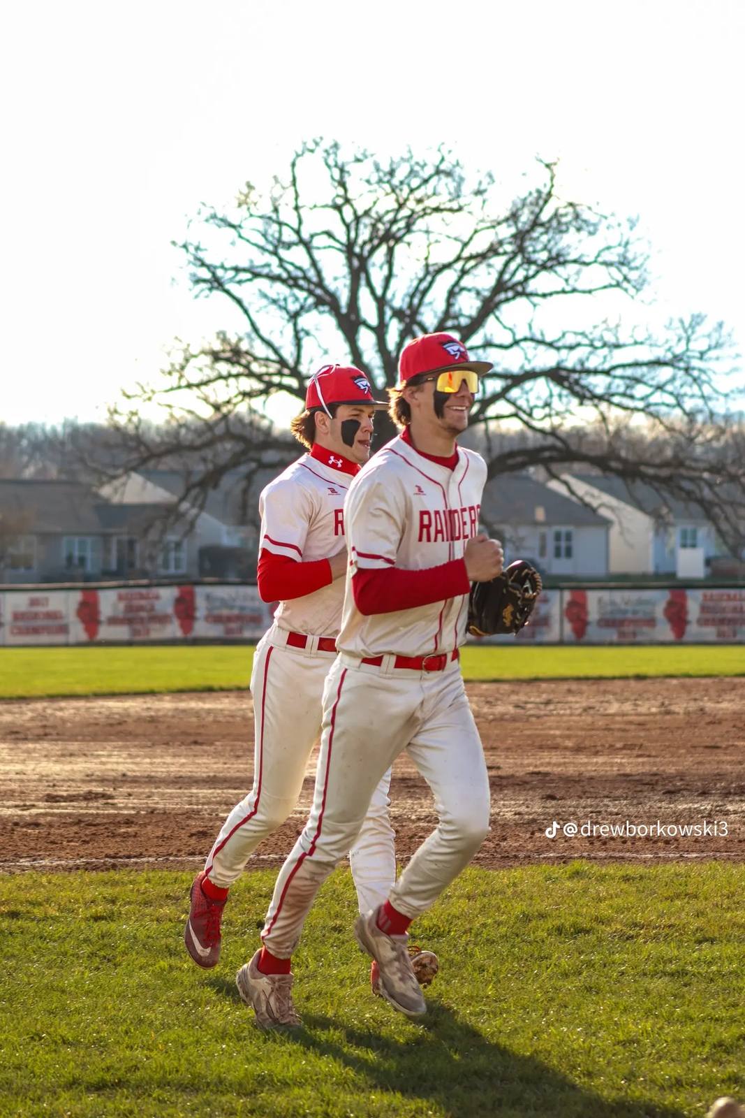 Two athletic young men wearing matching red and white baseball uniforms jog across a sunlit baseball field.