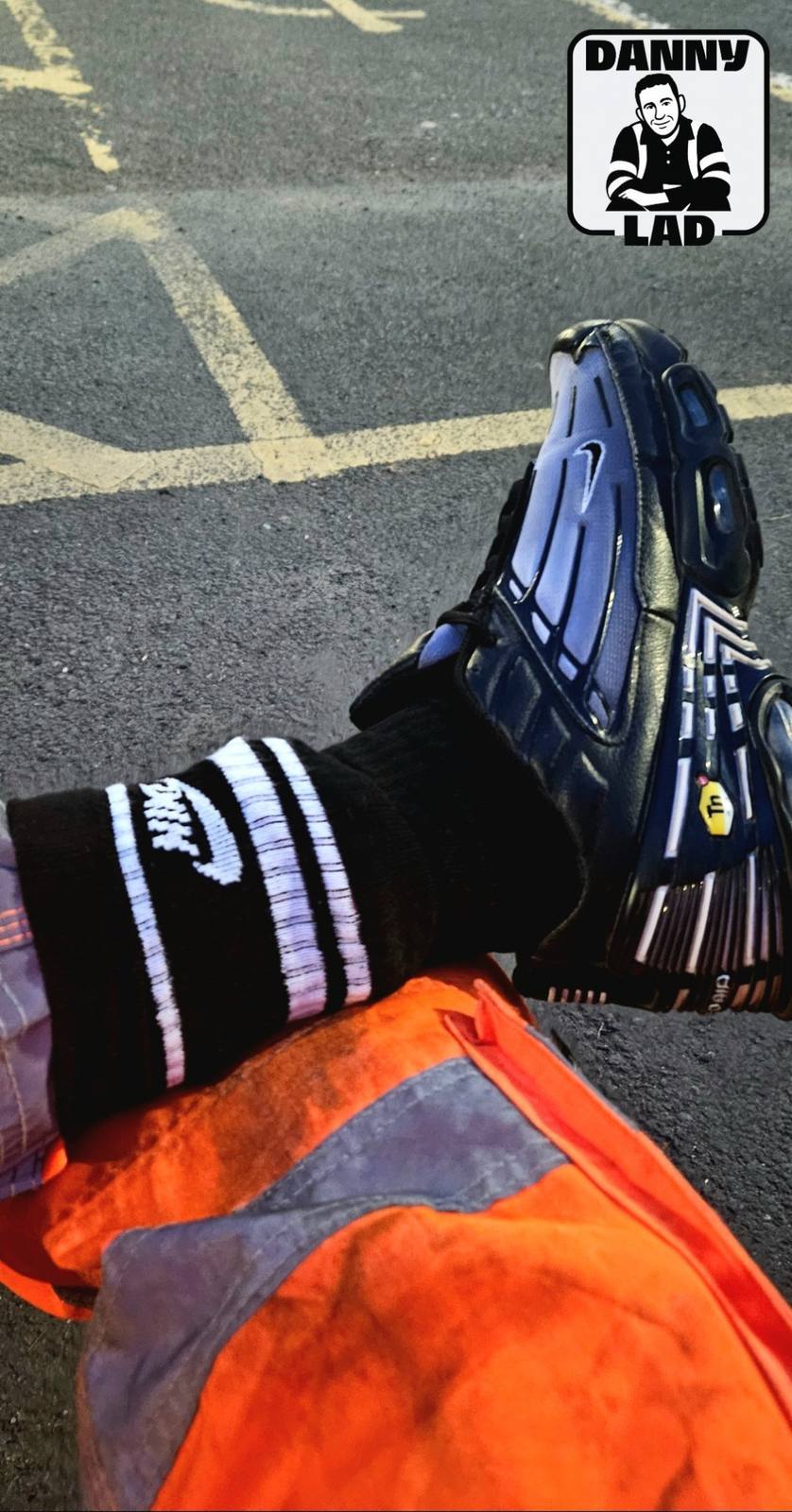 A seated person shows off their black Nike socks and blue TN sneakers while wearing hi-vis orange work trousers in an outdoor setting.