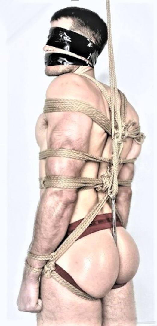 A muscular man is secured in intricate rope bondage with a tape blindfold and gag, wearing a jockstrap.